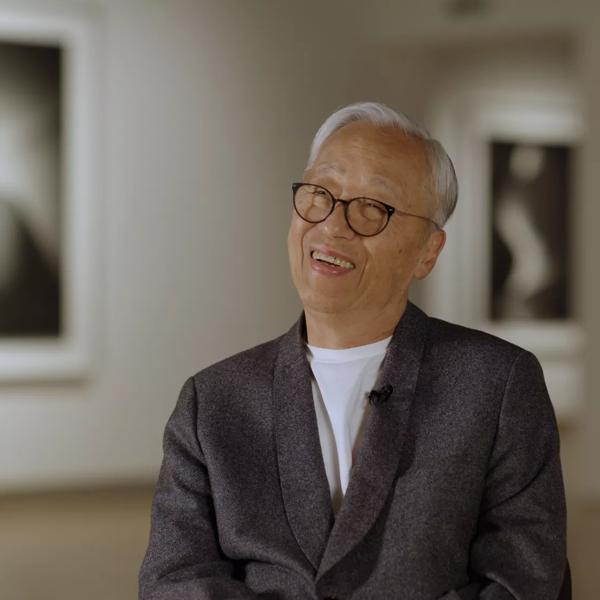 Hiroshi Sugimoto, an older Japanese man with white hair, wearing glasses and a grey suit jacket over a white t-shirt sits in the Hayward Gallery, behind him are two of his art works, part of the Hayward Gallery's Hiroshi Sugimoto exhibition