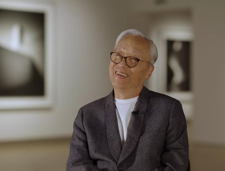 Hiroshi Sugimoto, an older Japanese man with white hair, wearing glasses and a grey suit jacket over a white t-shirt sits in the Hayward Gallery, behind him are two of his art works, part of the Hayward Gallery's Hiroshi Sugimoto exhibition