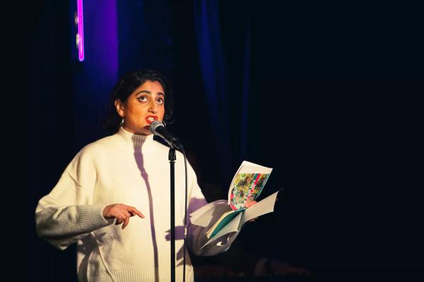 Shagufta K Iqbal reading aloud from a book of poetry with a microphone wearing a white jumper.