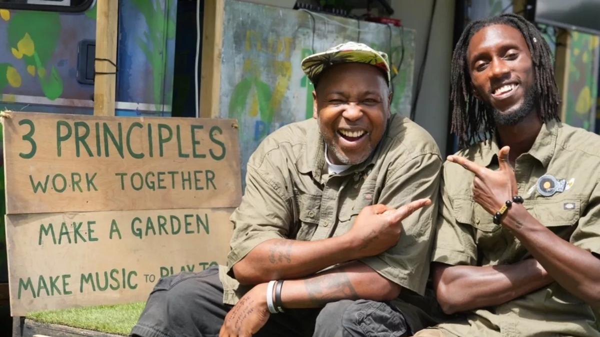 Two Black men wearing green shirts smiling and pointing at each other, next to sign which reads '3 Principles: Work Together, Make a Garden, Make Music to Play Together'