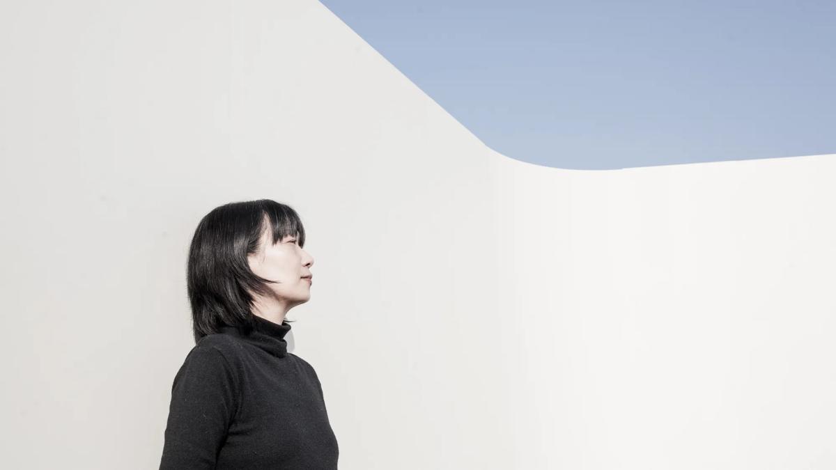 Han wears a black turtleneck and is pictured in side-profile. She's got cropped black hair and a fringe and is pictured outdoors standing against a white wall. Blue sky is visible in the top right corner of the image. 