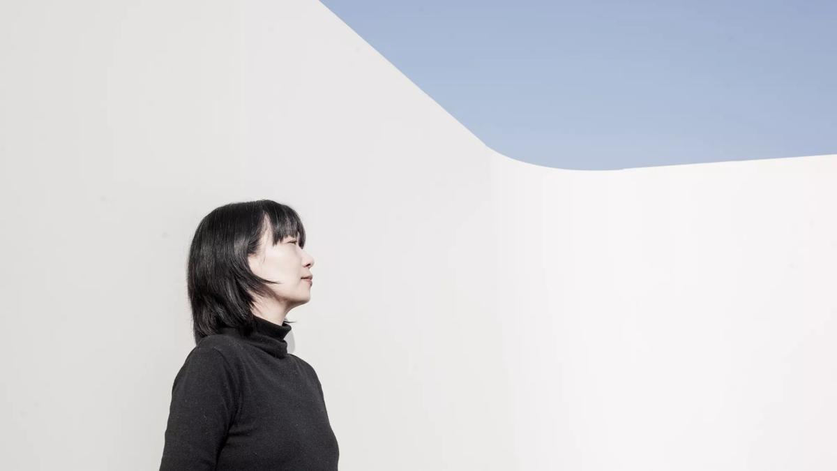 Han wears a black turtleneck and is pictured in side-profile. She's got cropped black hair and a fringe and is pictured outdoors standing against a white wall. Blue sky is visible in the top right corner of the image. 