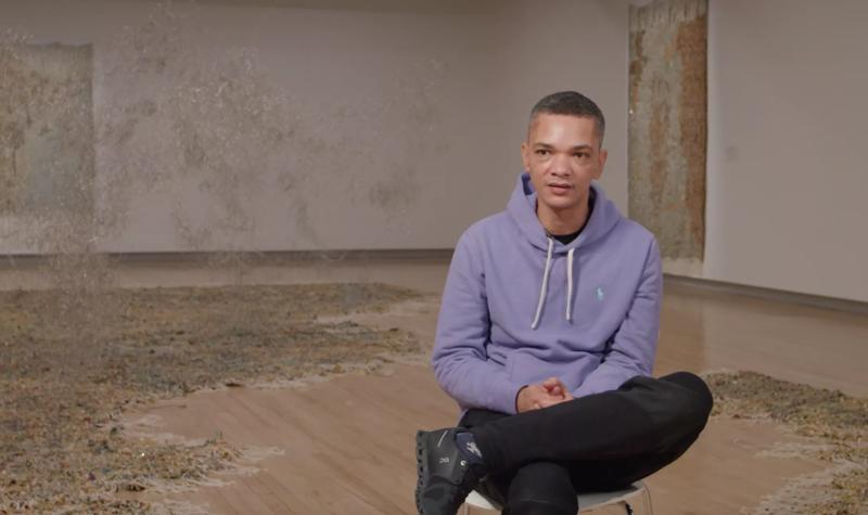 The artist Igshaan Adams sits cross-legged on a chair in the middle of his Hayward Gallery installation Kicking Dust as he talks to camera during an interview