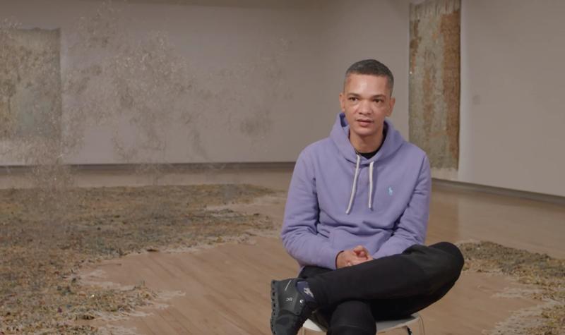 The artist Igshaan Adams sits cross-legged on a chair in the middle of his Hayward Gallery installation Kicking Dust as he talks to camera during an interview