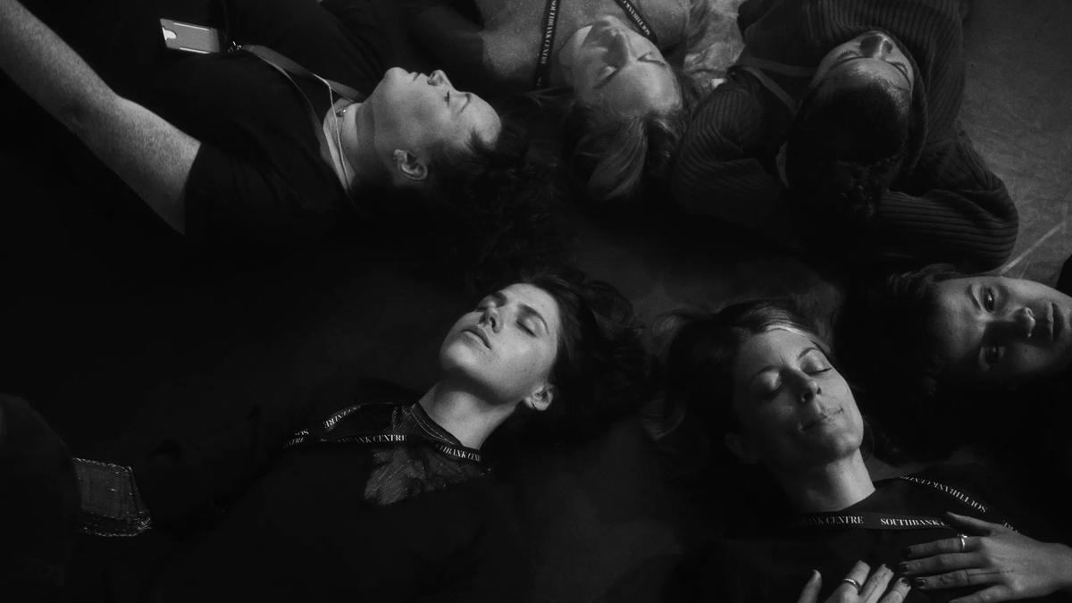 6 people lay on the floor with their eyes closed in black and white. 