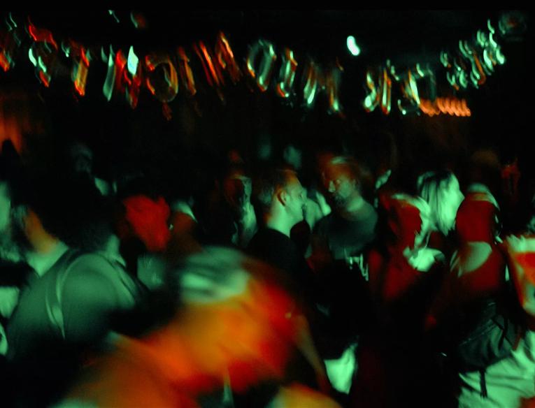 Crowd of people in a dark venue under green and red lighting with silver letter shape balloons hung above