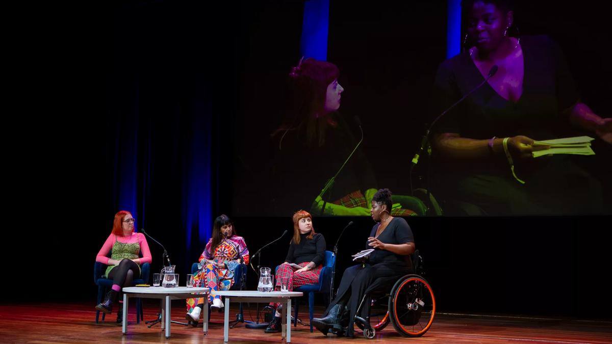 Four speakers on the Royal Festival Hall stage, the lady on the right is in a wheelchair. There is a projection still of two women behind them
