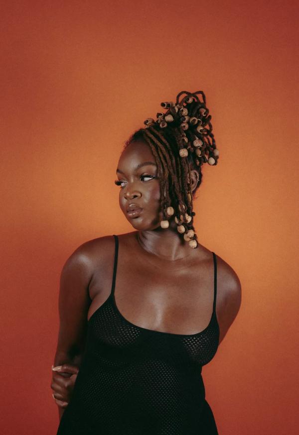 Artist Lizzie Berchie wears a black vest and stands in front of an orange background with wooden beads in her hair