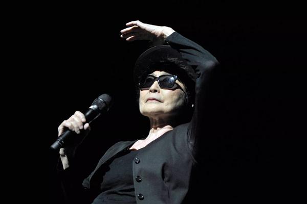 Yoko Ono on stage at her 2013 Meltdown Festival, she is wearing a black brimmed hat and sunglasses and holds a microphone in her right hand whilst her left had shields her eyes from the stage lights