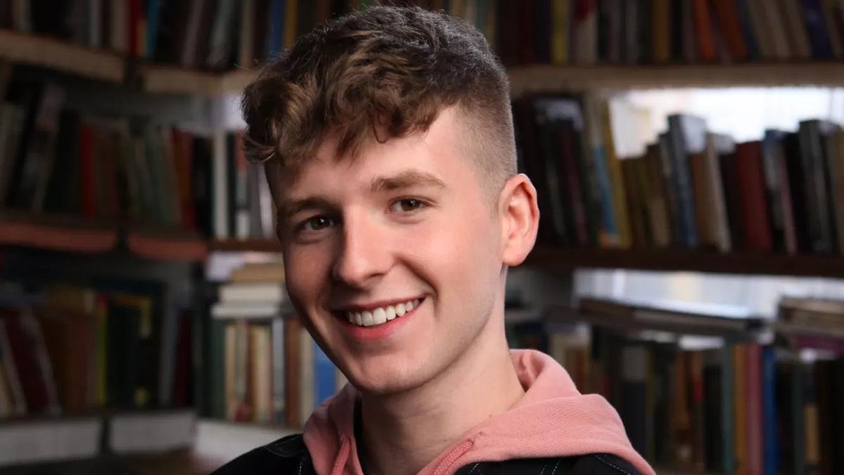 Portrait of YouTube personality Adam B. He stands amongst shelves of books smiling to the camera. He wears dark collared jacket with white seams and a salmon pink hoodie. 