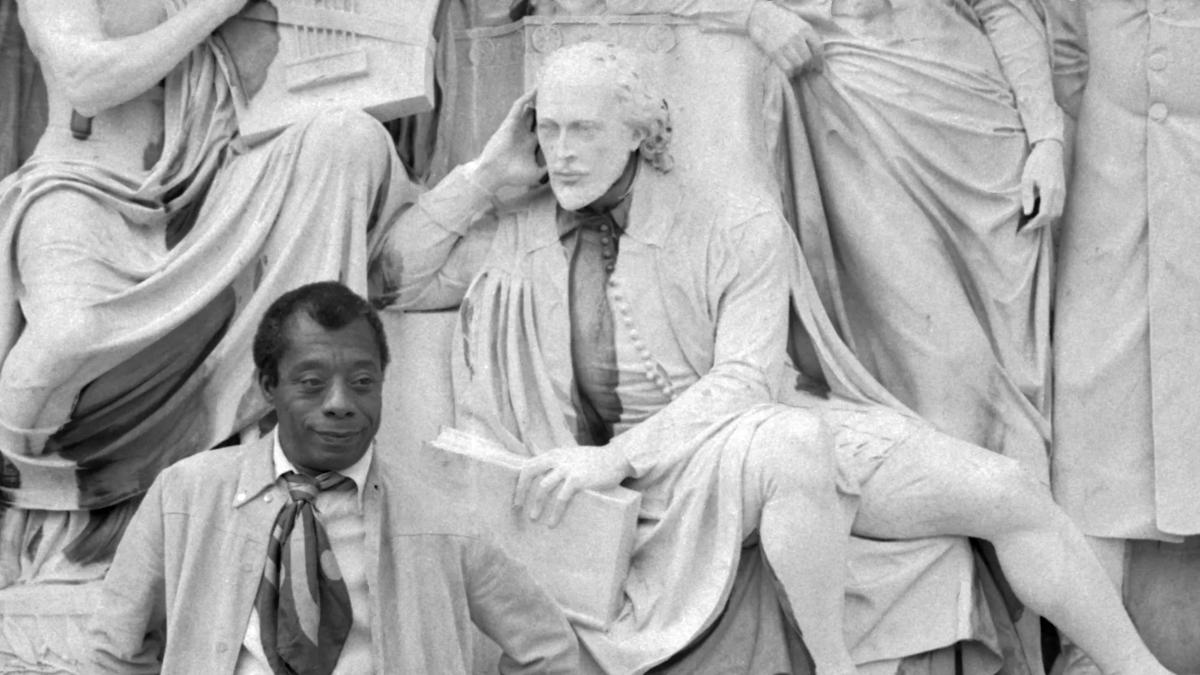 Black and white photo of author James Baldwin standing in front of a statue