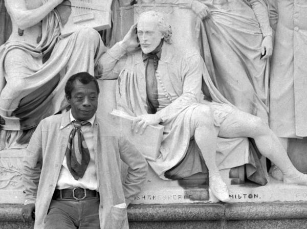 Black and white photo of author James Baldwin standing in front of a statue