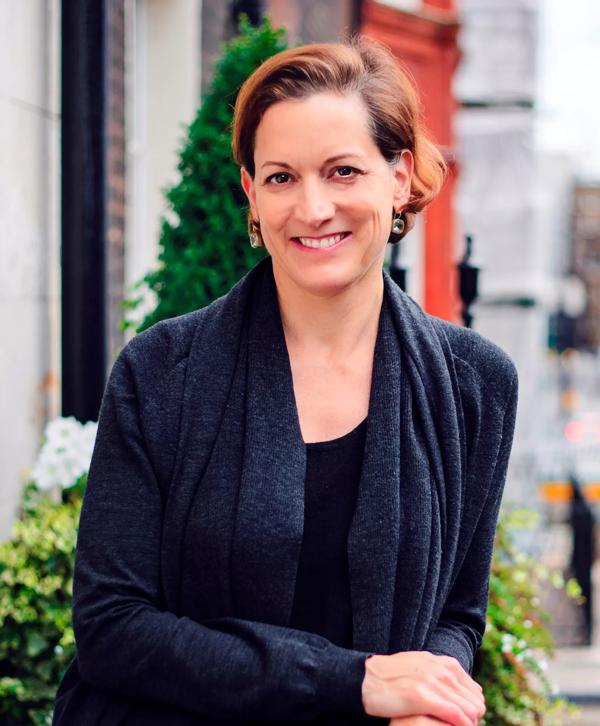 Portrait of Anne Applebaum, smiling as she stands outside. She wears a dark grey cardigan and black top