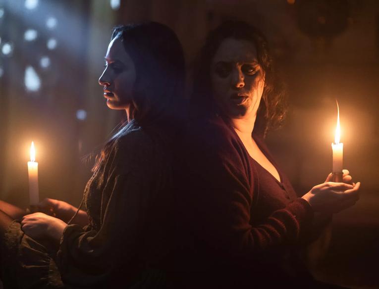 Two women sit back to back in the dark. They hold candles which light their faces.