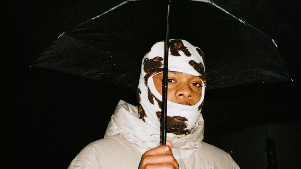 Musician Tay Jordan sporting a white and brown baclava under a black umbrella wearing a cream coloured puffer jacket