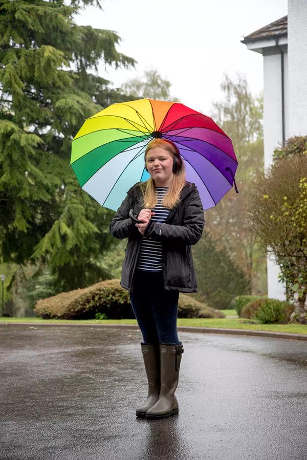 Rebecca stands outside a house, wearing headphones and holding a rainbow coloured umbrella