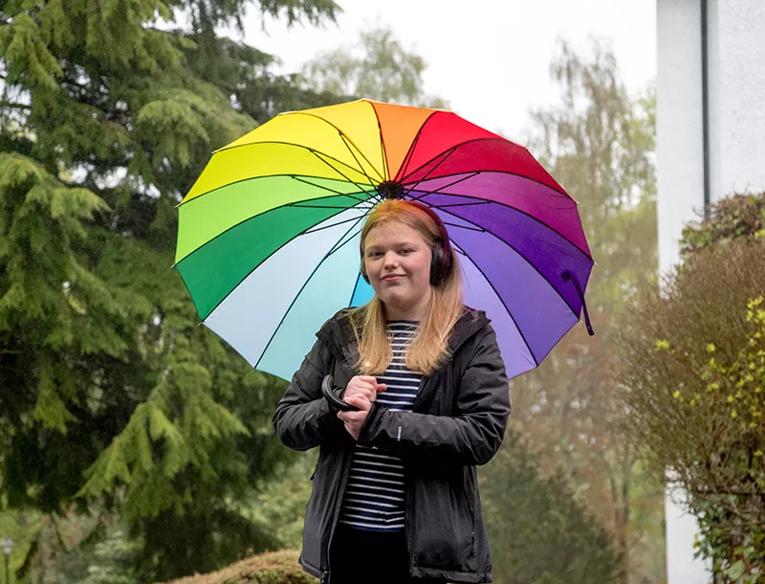 Rebecca stands outside a house, wearing headphones and holding a rainbow coloured umbrella