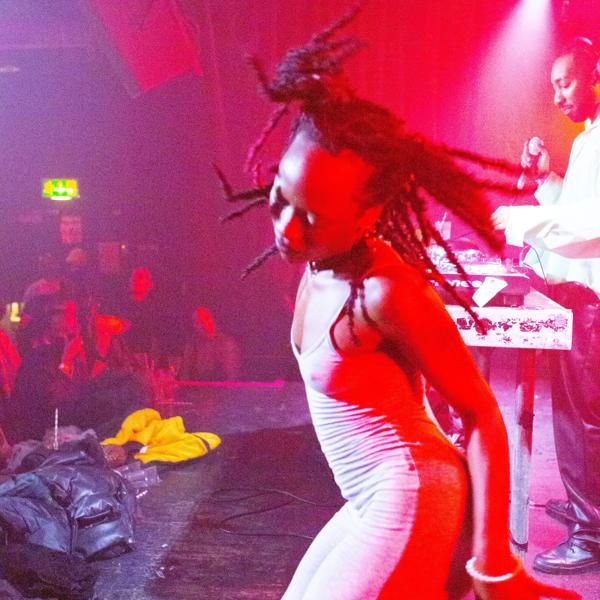A black artist dancing on the stage. In the back view is a DJ and some crowd of people  
