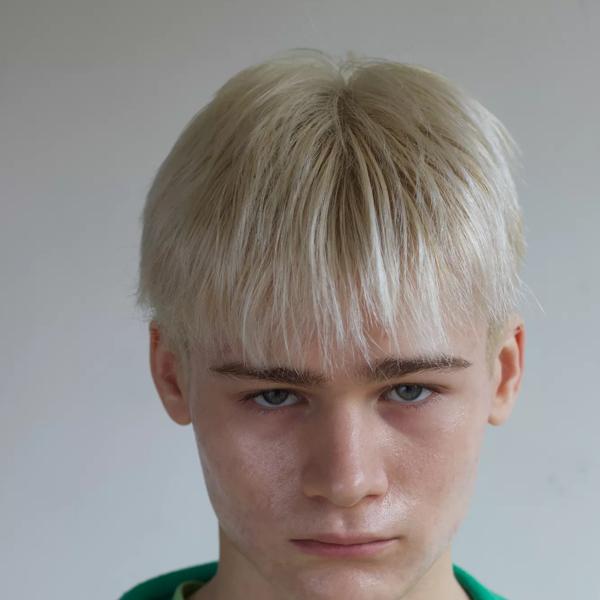 Abe has bleached blonde hair and blue eyes. He has freckles and looks directly into camera. He wears a light green t-shirt and dark green hoodie. 