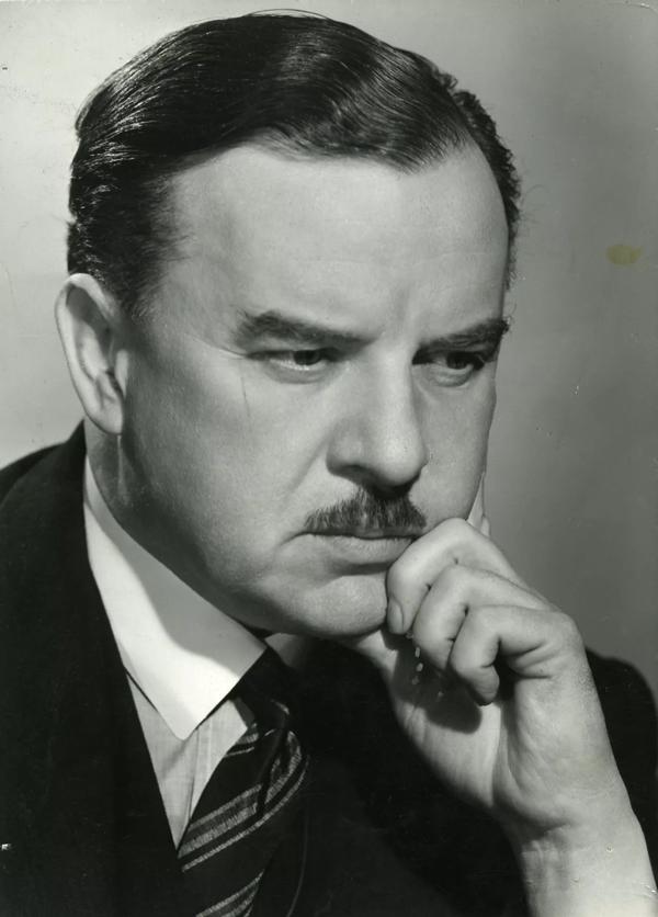 A 1950s headshot of Sir George Thalben-Ball; his dark hair is laquered and he has a small moustache; he wears a suit and tie and is resting his chin on his left hand