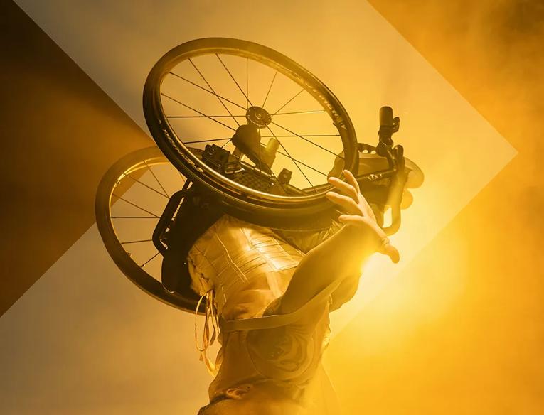 A dancer in a wheelchair leans upside down on the head with the wheelchair in the air in front of a bright yellow light.