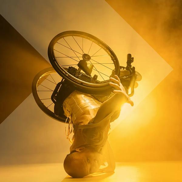 A dancer in a wheelchair leans upside down on the head with the wheelchair in the air in front of a bright yellow light.