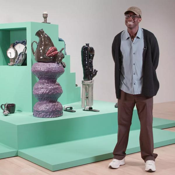 Sculptor Woody De Othello, wearing trainers and a baseball cap, stands in front of his ceramic works which feature in Strange Clay at Hayward Gallery