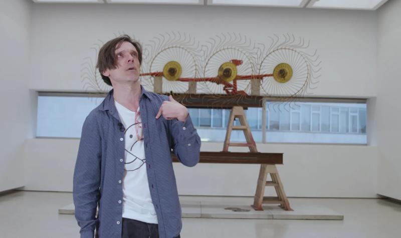 The artist Jeremy Deller wearing an open blue shirt over a white t-shirt stands inside the Hayward Gallery in front of a work by Mike Nelson, part of the Mike Nelson: Extinction Beckons exhibition at Hayward Gallery