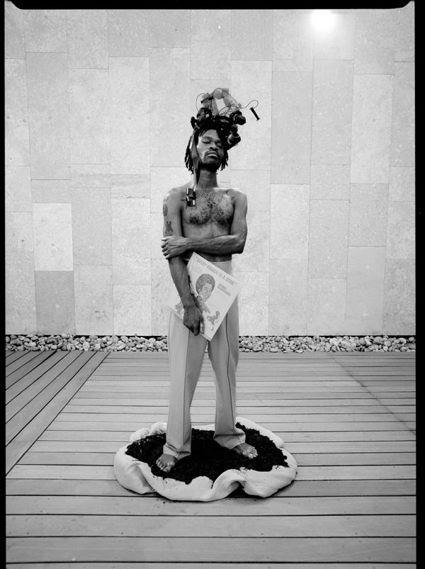 A topless Black man with an elaborate headress stands holding his right elbow with his left arm. He has a vinyl record sleeve in his right hand