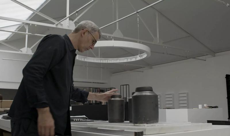 Edmund de Waal, a white man with grey hair, wearing glasses and a dark shirt stands in his studio a white walled and ceilinged space; he touches a ceramic vessel on a counter top
