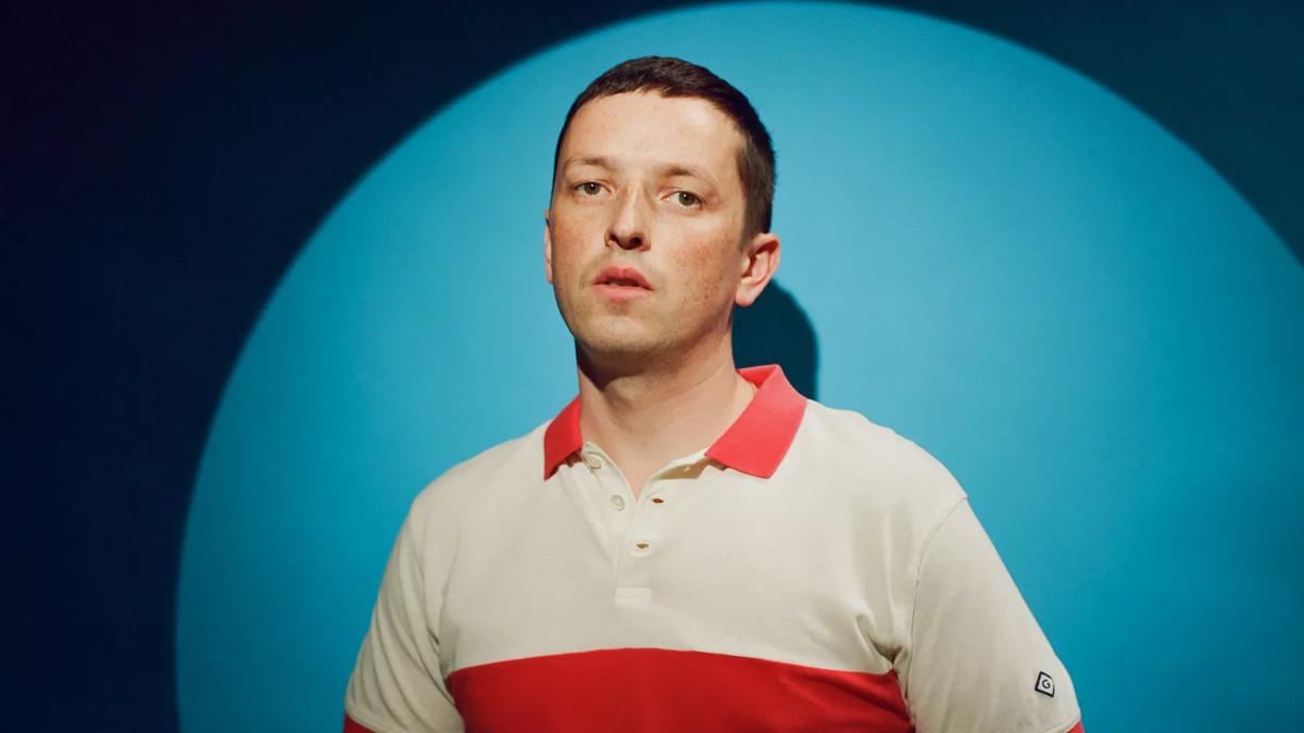 Portrait of artist Totally Extinct Enormous Dinosaurs in a white and red polo shirt in front of a blue background lit by a spotlight.