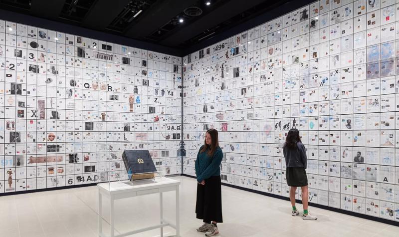 Installation view of a large encyclopaedia, in a room with walls covered in paper clippings.