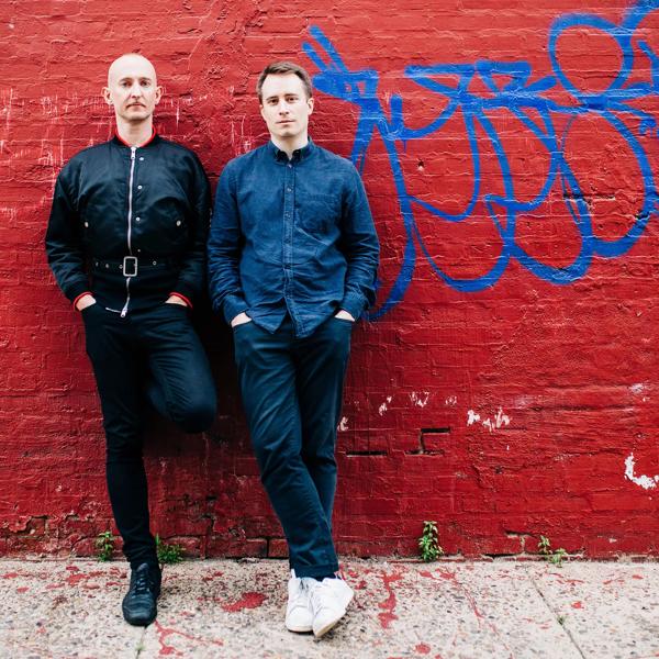 Philip Venables and Ted Huffman stand together outside, leaning against a brick wall which has been painted red and has the remnants of graffiti on it. Philip is a white bald man wearing a leather jacket, Ted has short brown hair and wears a denim shirt
