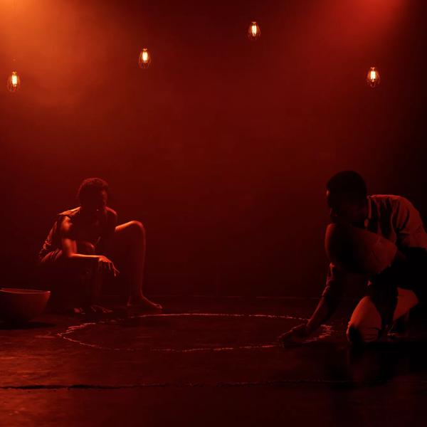 The image features two black performers crouched down on a black floor.   The lighting is dark and atmospheric. There is a golden orange light shining down on them.   The female performer is kneeling in the background, pointing to the middle of a circle made from lentils.  The male performer is pouring lentils in a circle from a wooden bowl. 