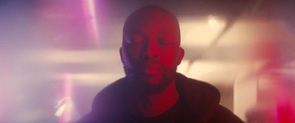 Head shot of a person with eyes closed surrounded by pink hazy lighting. They have dark skin, a shaved head and facial hair.
