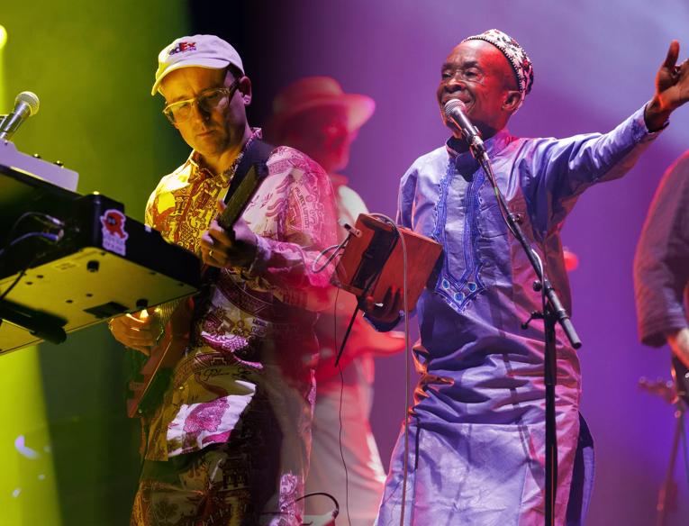 A composite image from Hot Congotronics performance at Grace Jones Meltdown that feature four members of the band, including Hot Chip's keyboardist a white man who wears a white baseball hat, a vocalist from Kasai Allstars, a black man who wears a long sleeve Dashiki, and in the background two male guitarists, each white men wearing wide brimmed hats are just visible but out of focus