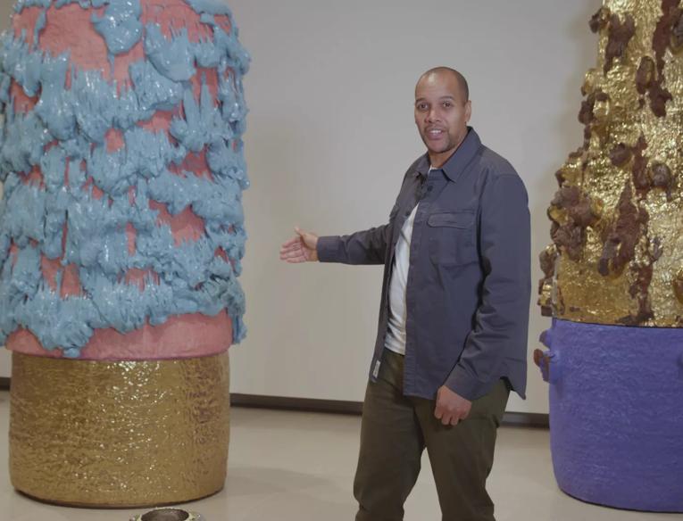 Ceramicist Rich Miller in Hayward Gallery, he wears an open blue overshirt over a white t-shirt. He is gesturing at the sculptural installation behind him, which is a huge clay sculpture in bright pink, with bright blue clay seeming to fall from it