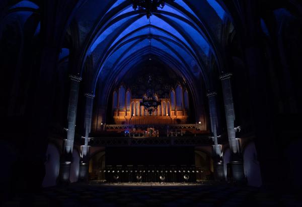 A grand organ under a big stone arch, lit up with yellow and blue light, and with several speakers attached.
