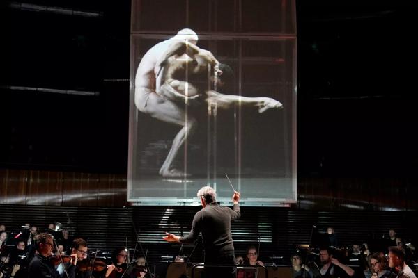 An orchestra with a screen suspended above, displaying dancers.