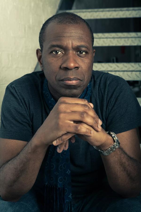 Portrait of Clive Myrie sitting on stairs with his hands clasped in front of his chest, wearing a black T-shirt