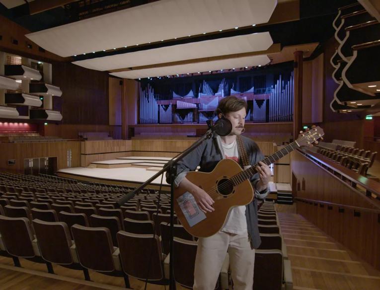 Vincent Neff of Django Django plays guitar inside an empty Royal Festival Hall. He is standing at a microphone stand among the auditorium seats, the stage and organ and boxes can be seen behind hime