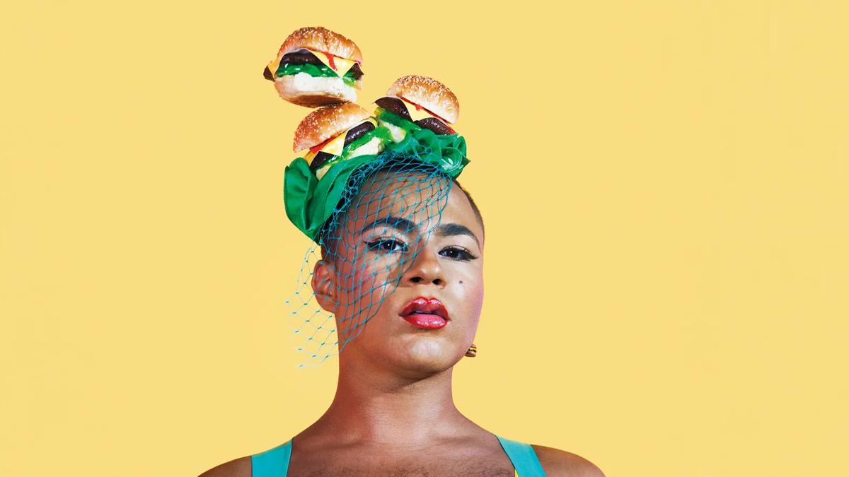 Performance artist Travis Alabanza wearing a fascinator headpiece, a layer of 3 cheeseburgers on top  