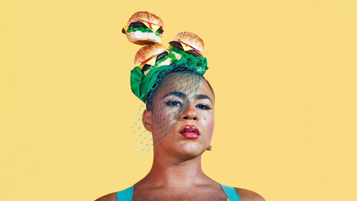 Performance artist Travis Alabanza wearing a fascinator headpiece, a layer of 3 cheeseburgers on top  