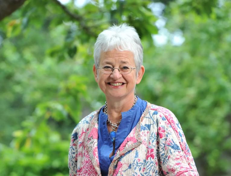 Jacqueline Wilson standing in front of green trees, wearing a flowery jacket, blue shirt and black trousers.