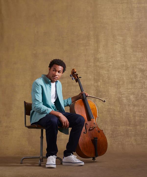 Cellist Sheku Kanneh-Mason in a denim shirt casually seated with his cello