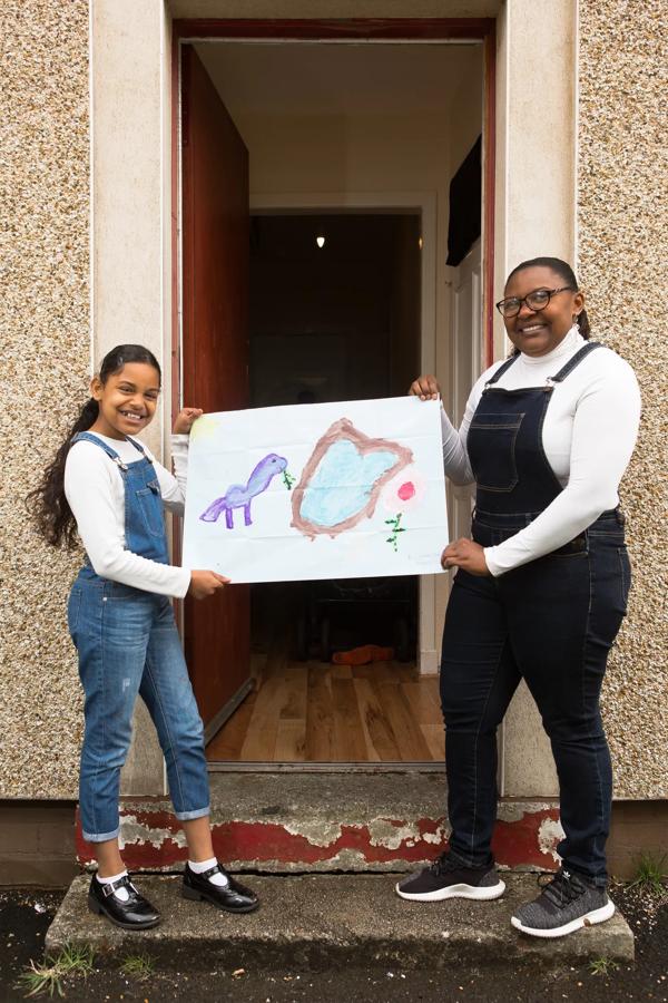 Art by Post participants Mary and her daughter Simmi stand on their front step jointly holding a piece of art work produced as part of Art by Post