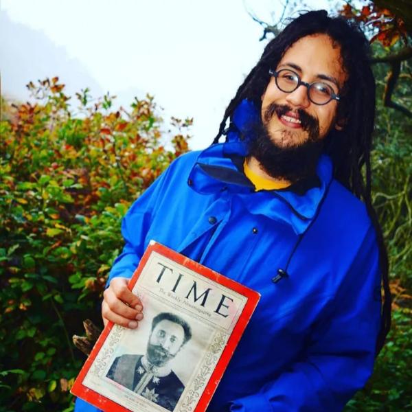 Ras Benji wearing a blue rain coat holding a vintage Time magazine in a garden