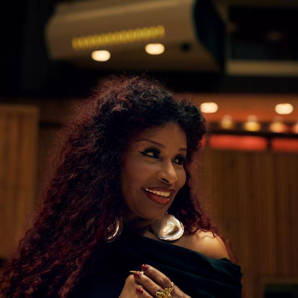 Head shot of Chaka Khan in the Royal Festival Hall, looking to the side and smiling with red-tinted curly hair and large gold earrings.