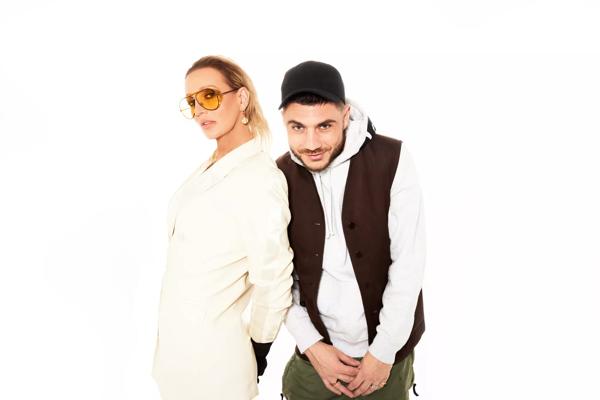 Two members of Panty Soaker Sound System stand against a white background. The person on the left wears a white coat and glasses and the person on the right wears a white hoodie, black waistcoat and black baseball cap.