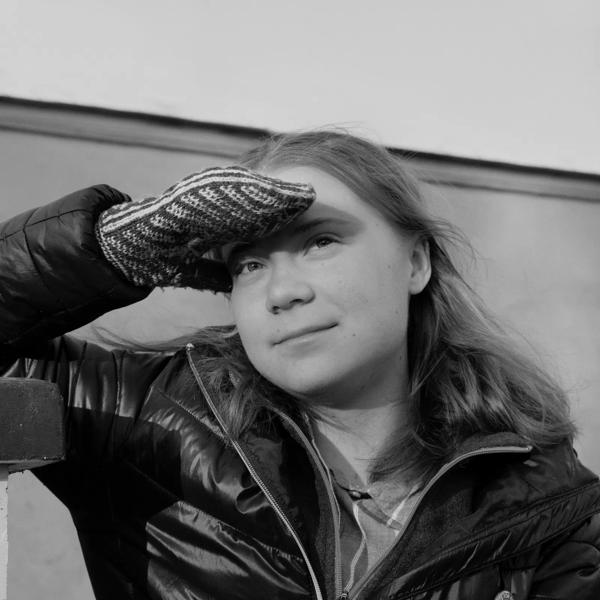 Black and white portrait of activist Greta Thunberg. She sits in a puffer jacket shielding her eyes from the sun in knitted mittens and is smiling slightly at the camera. 